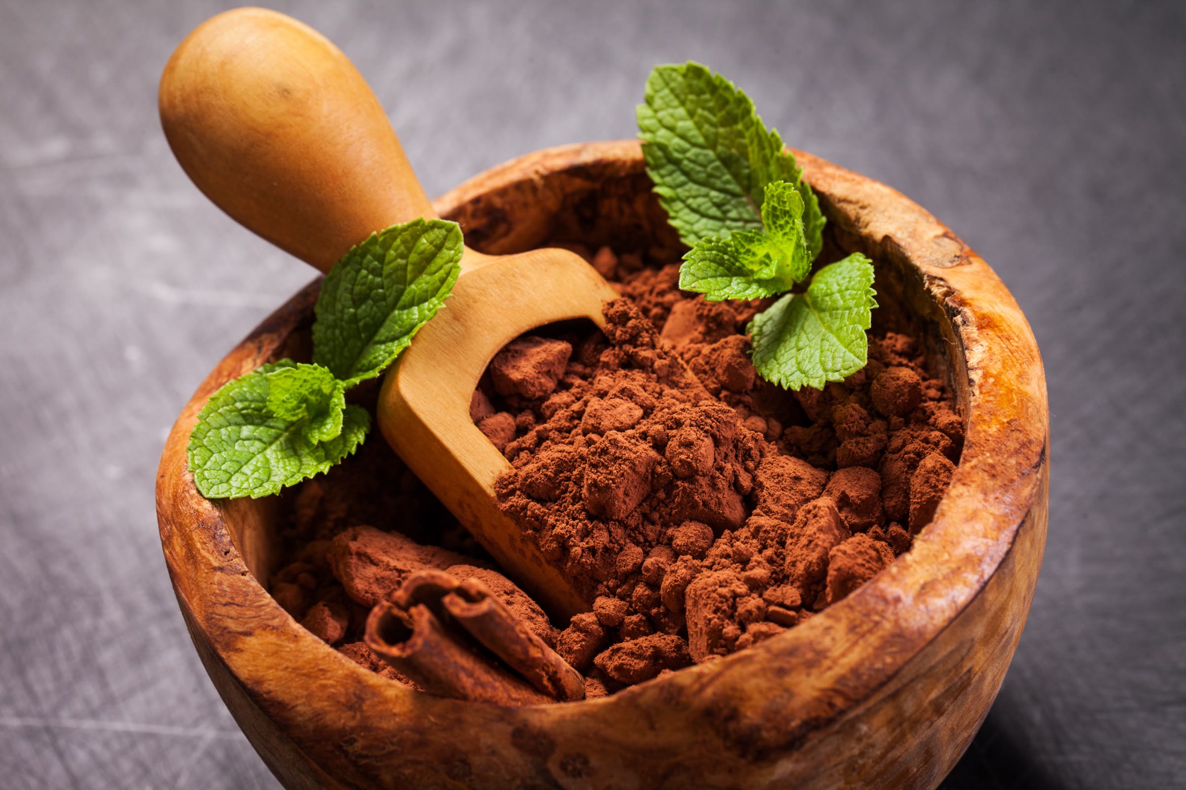 A wooden scoop and fresh mint sprigs in a bowl of cocoa powder